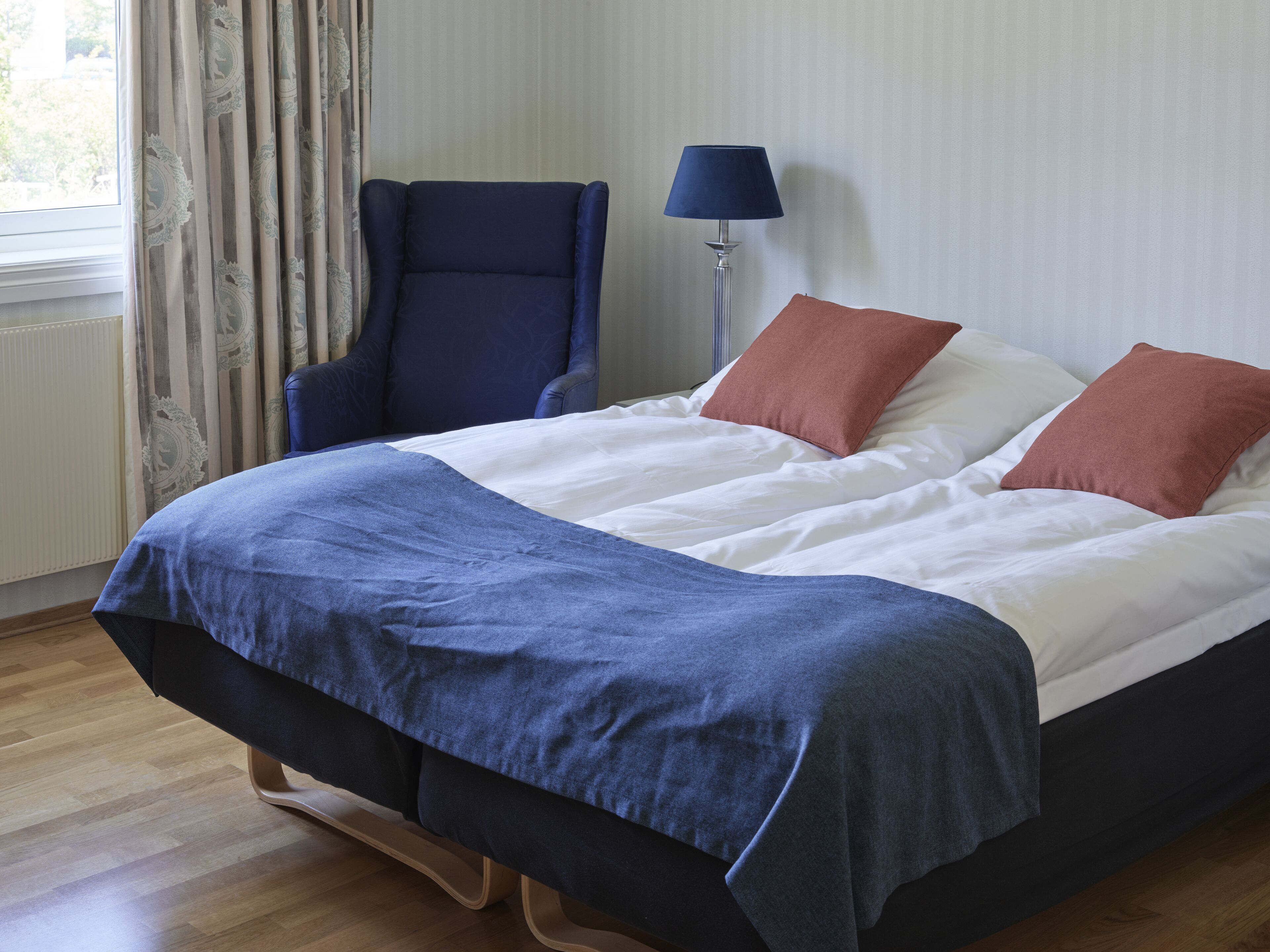 Ein ordentlich bezogenes Bett mit einer blauen und weißen Bettdecke, zwei braunen Kissen und einem blauen Sessel daneben.