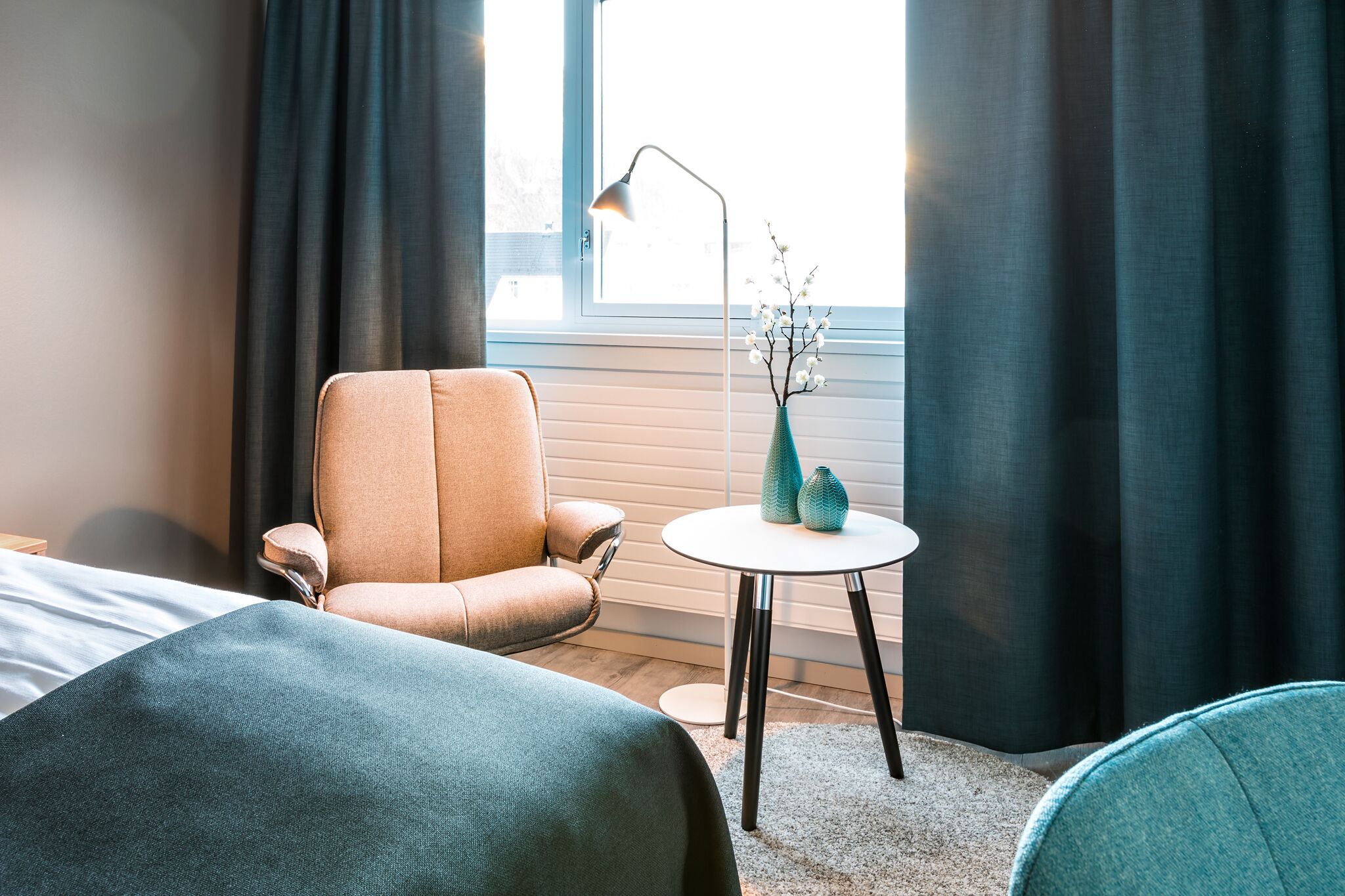 A hotel room with a bed, a chair, a small table with vases, and a window with curtains.