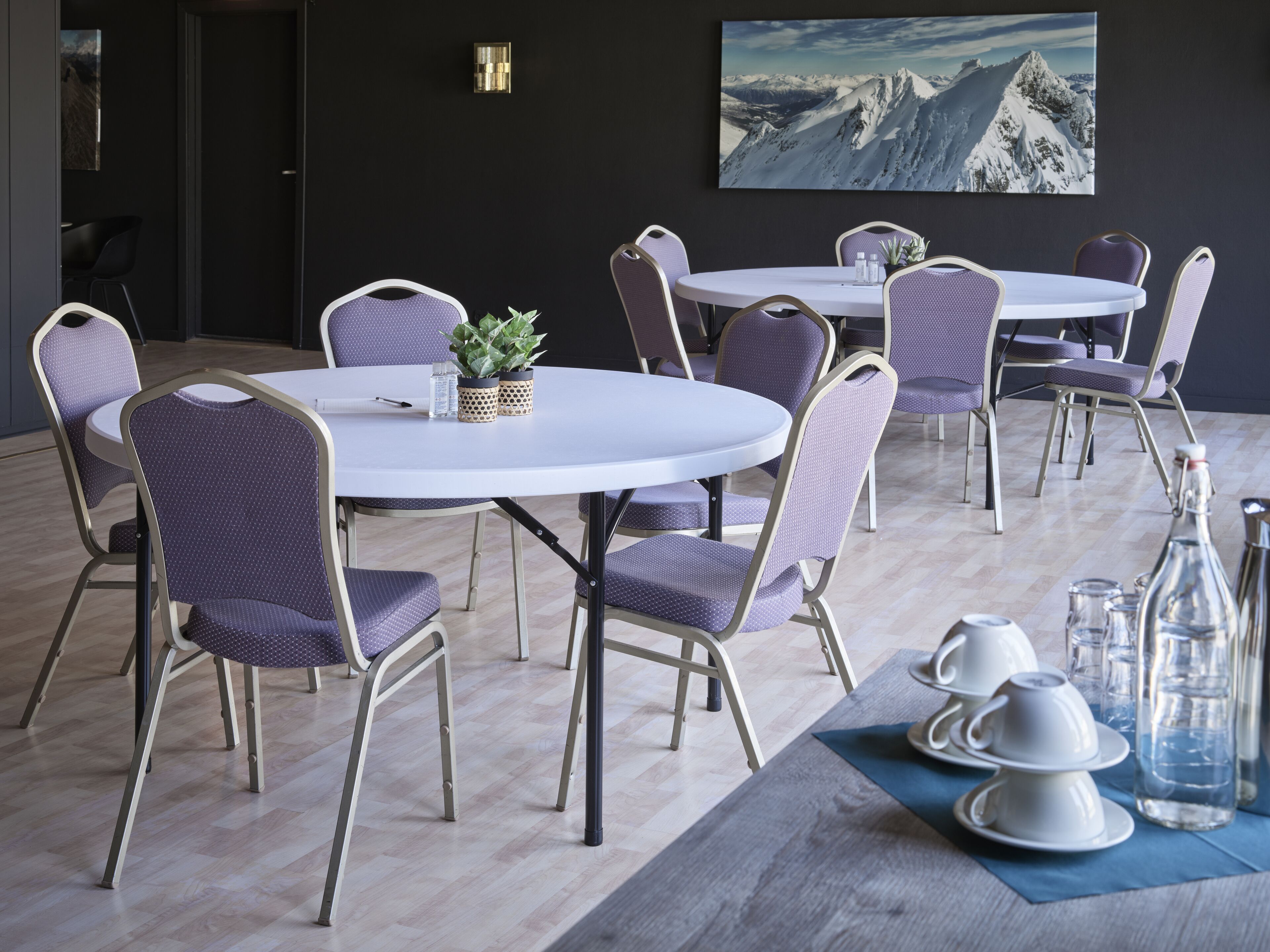 Een moderne eetruimte met ronde tafels en paarse beklede stoelen, met een schilderij van een berglandschap aan de muur.