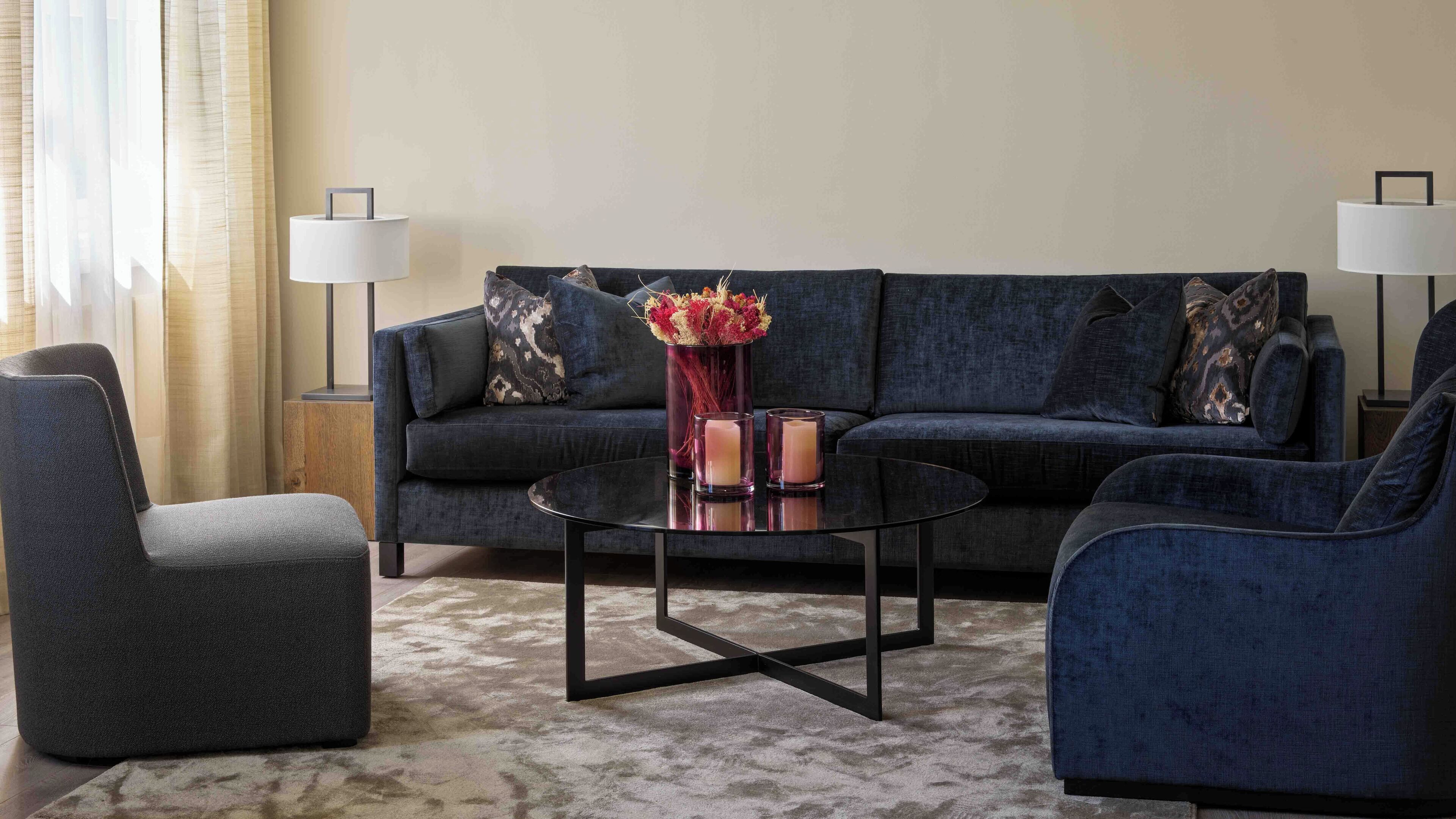 A modern living room with a dark blue sofa, two armchairs, a glass coffee table, and a vase with flowers. There are two floor lamps and a side table with a lamp.