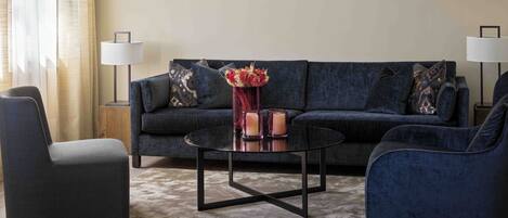 A modern living room with a dark blue sofa, two armchairs, a glass coffee table, and a vase with flowers. There are two floor lamps and a side table with a lamp.