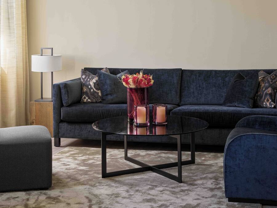 Una sala de estar moderna con un sofá azul oscuro, dos sillones, una mesa de centro de vidrio y un jarrón con flores. Hay dos lámparas de pie y una mesita de noche con lámpara.