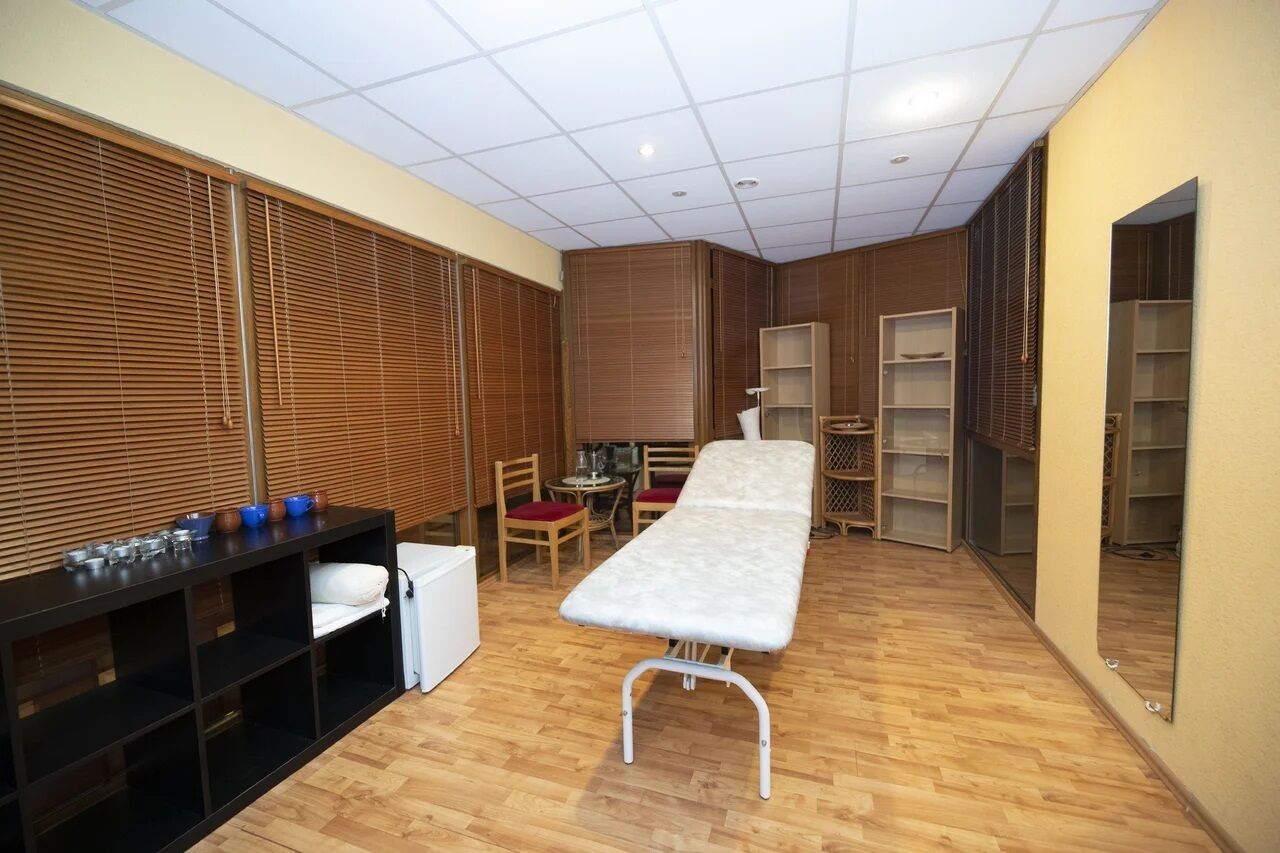 Una habitación con piso de madera, una cama, una pequeña mesa con sillas y un estante negro con diversos objetos.