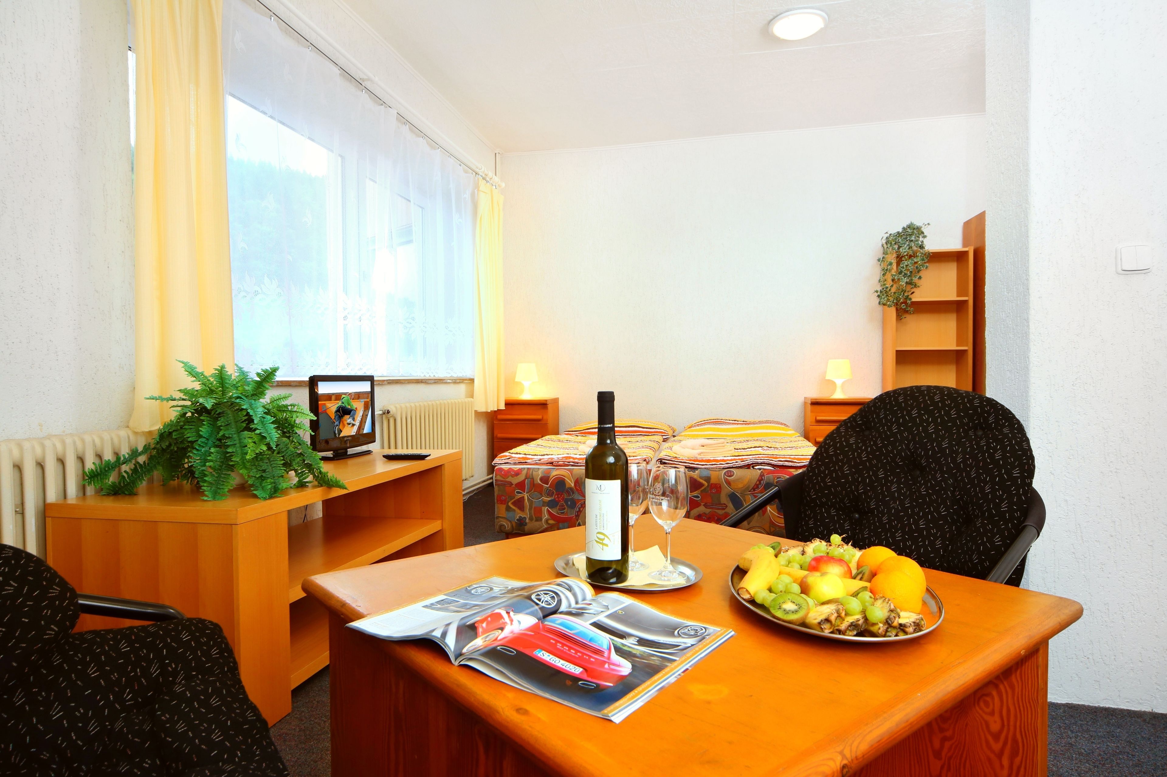 A hotel room with a wooden desk, a dining table with a fruit plate, a bottle of wine, and a magazine. There is a window with curtains, a radiator, and a plant on the wall.