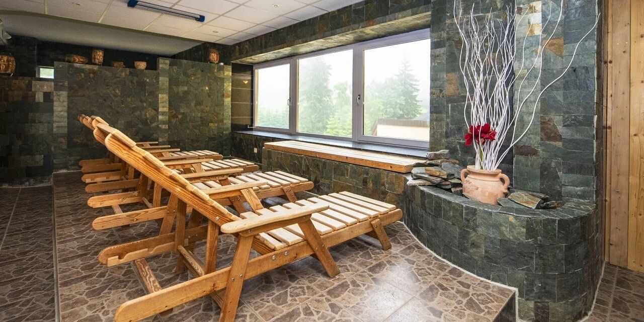 A room with wooden benches arranged in rows, a stone wall, a window with a view, and a vase with branches.