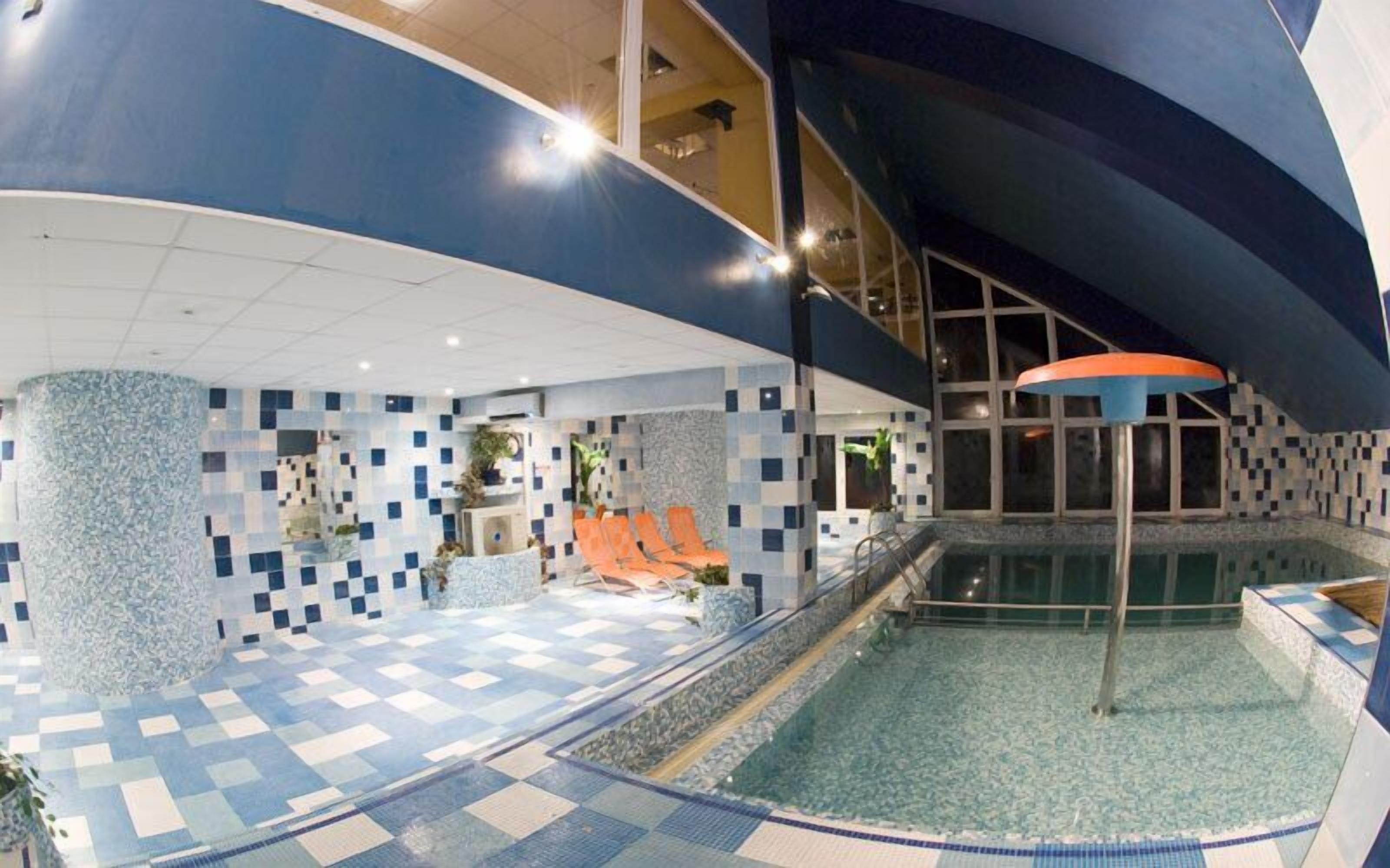 Un área de piscina interior moderna con suelo a cuadros, zona de estar con sillas anaranjadas y un ventanal grande con vistas a la piscina.