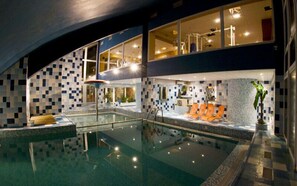 A modern indoor pool area with clear glass walls, a seating area with orange loungers, and a tiled wall design. - Hotel Fit & Fun (Harrachov)