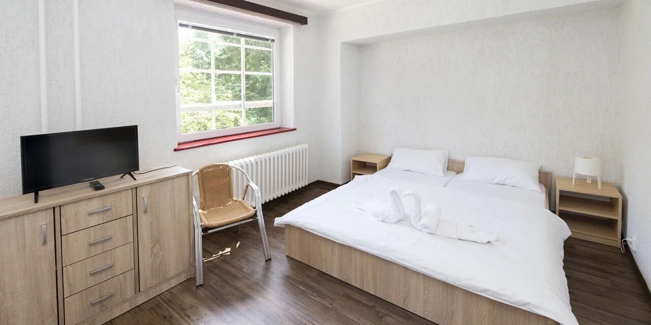 Un dormitorio con cama, televisor sobre un mueble de madera, una silla, ventana y mesita de noche.