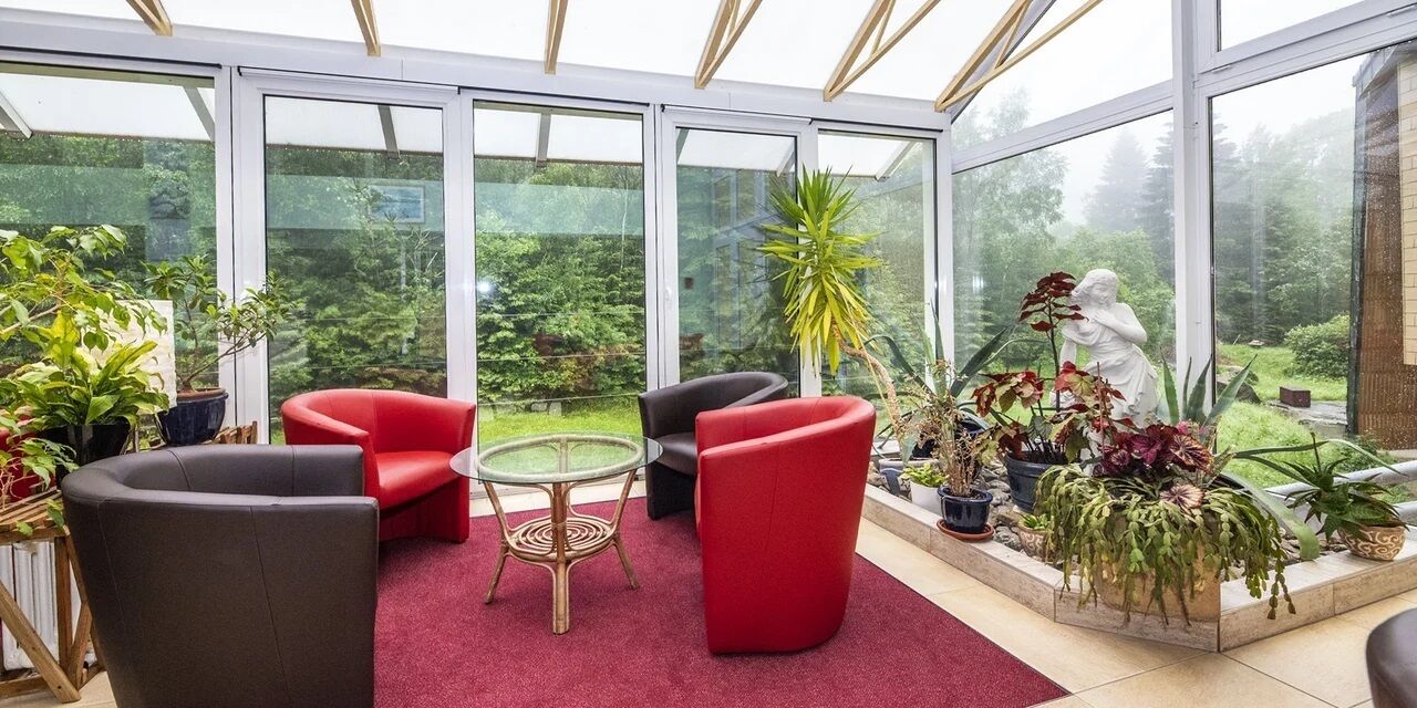 A conservatory with glass walls, red and black chairs, a small round table, and various potted plants.