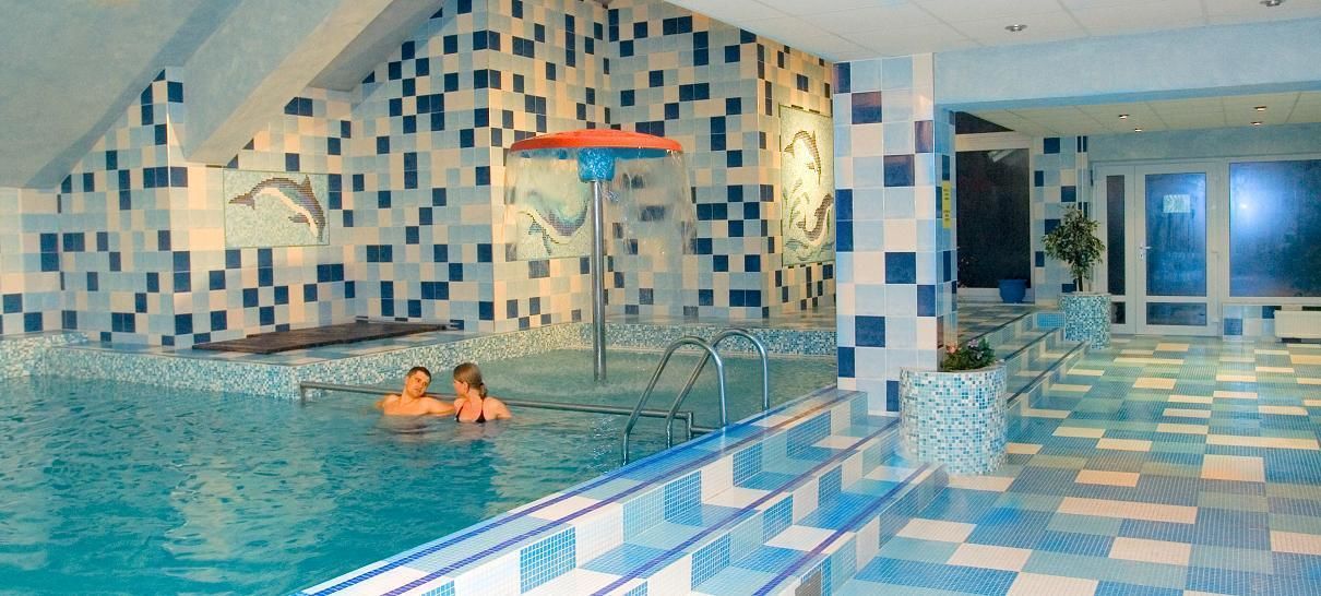 Ein Hallenbad mit gefliesten Wänden und einem Schachbrettmuster-Boden, einem Sprungbrett und einer Fontäne.