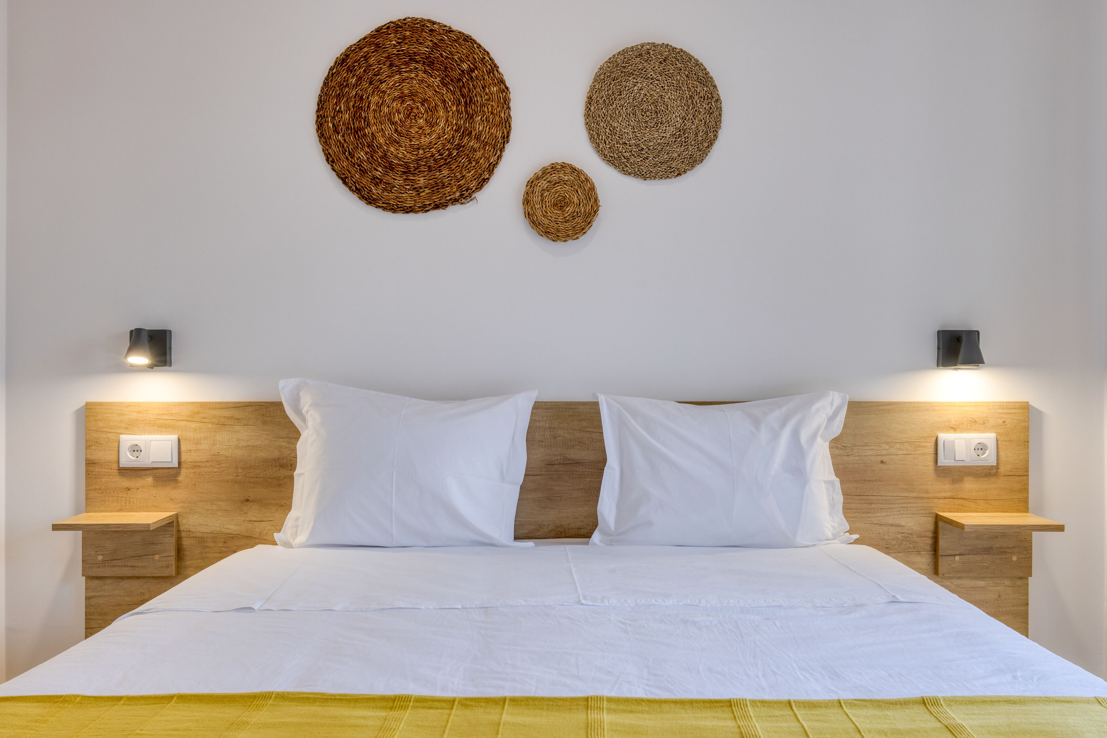 Uma cama bem arrumada com lençóis brancos, duas mesas de cabeceira com abajures e três peças de arte de parede feitas de fibras.