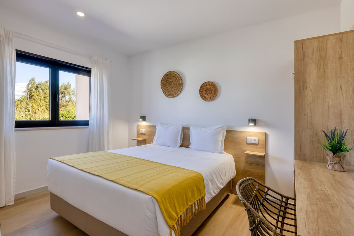 A modern hotel room with a large bed, a wooden headboard, two wall-mounted lights, a small table with a plant, and a view of trees through the window.