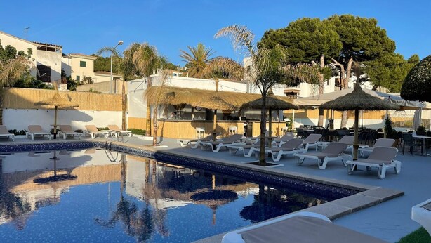 A pool area with lounge chairs and umbrellas, surrounded by buildings and trees. - Hotel Chevy & Suites (Capdepera)