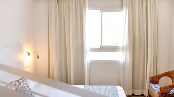 A hotel room with a bed, a window with curtains, a wall-mounted light, and a wicker chair.