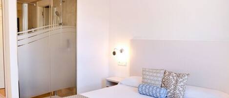 Un dormitorio con cama, mesitas de noche, luz instalada en la pared y un baño con ducha.