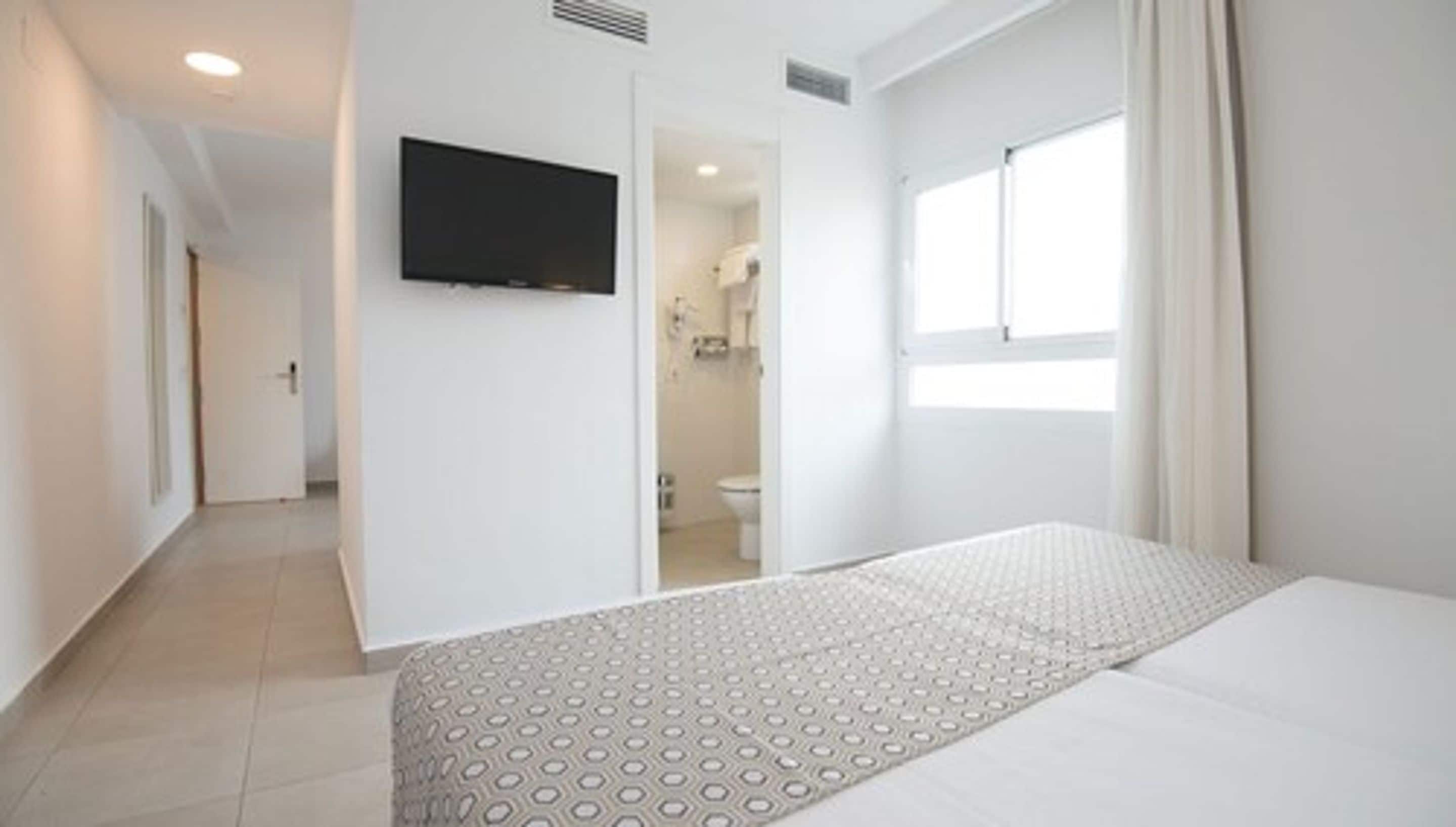 A hotel room with a bed, a TV mounted on the wall, a bathroom visible through an open door, and a window with curtains.