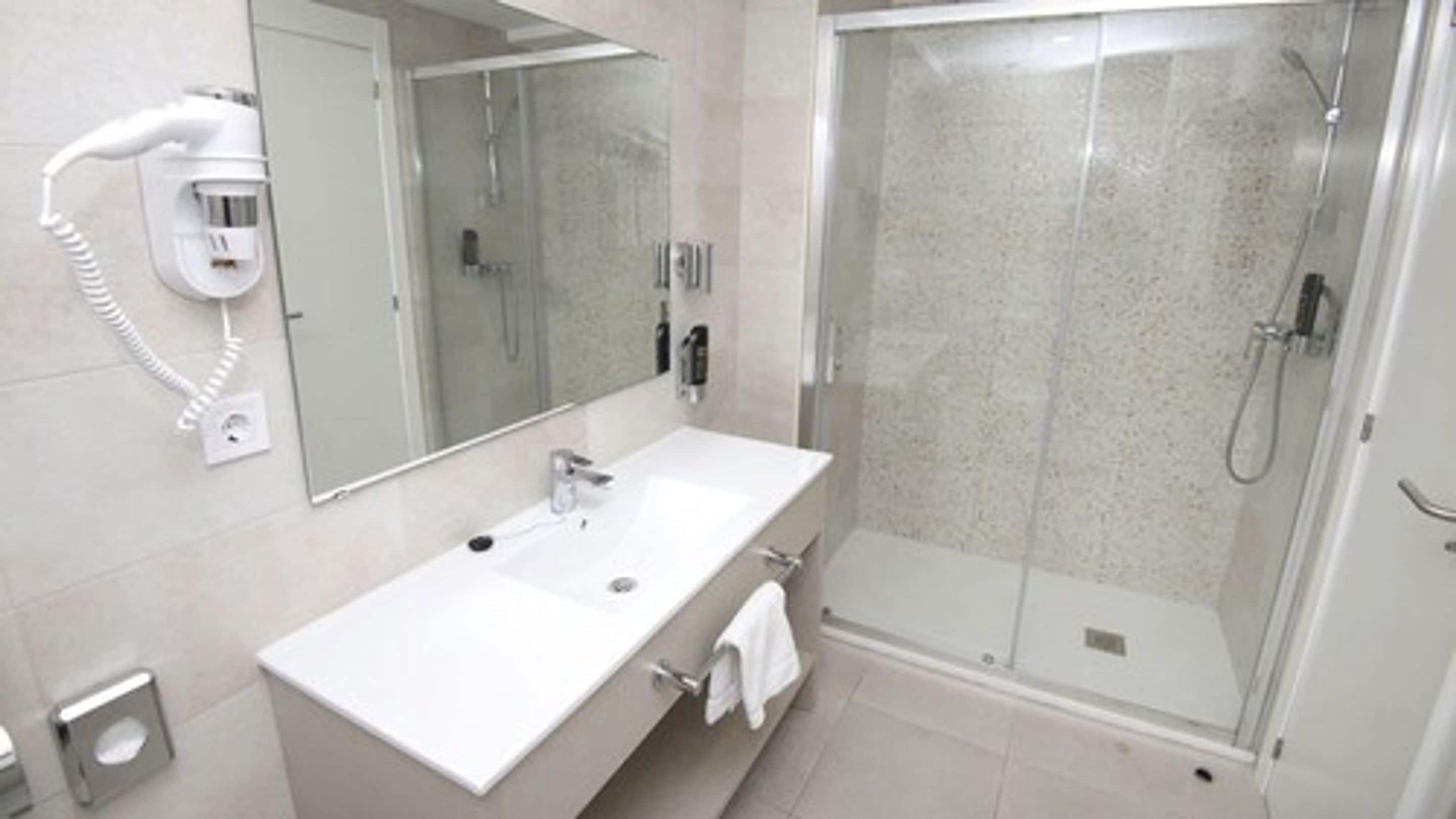 A modern bathroom with a white sink, a large mirror, a hairdryer, and a glass-enclosed shower.