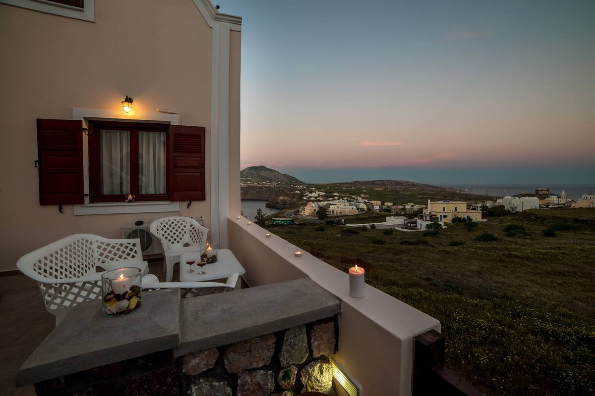A balcony with a table and chairs, a candle, and a view of a coastal village.