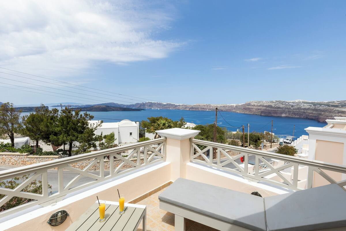 A balcony with a table and a drink, overlooking a coastal landscape with buildings and a clear sky.