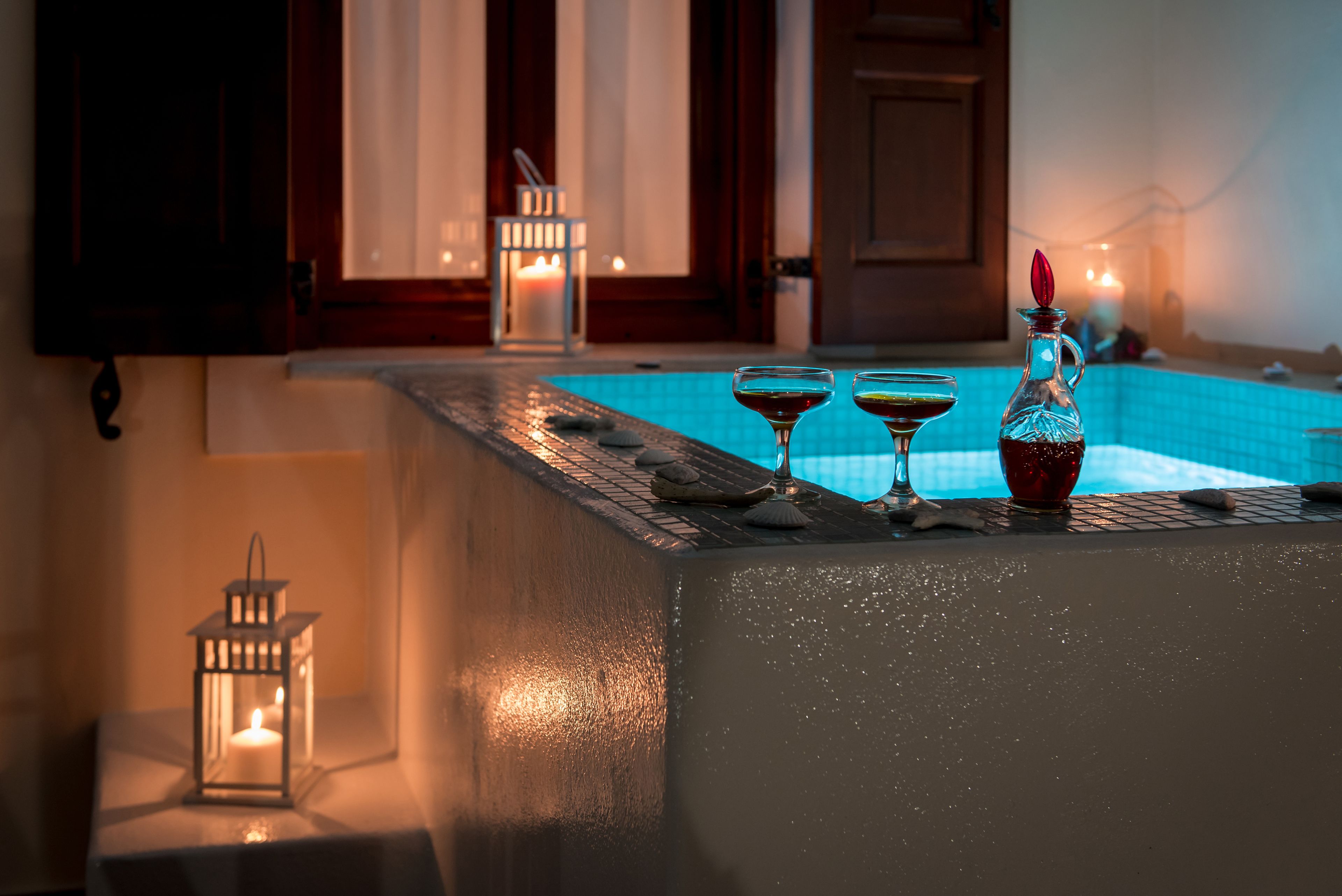 A poolside area with a tiled edge, two glasses of drink, a bottle, and a lit candle on a table.