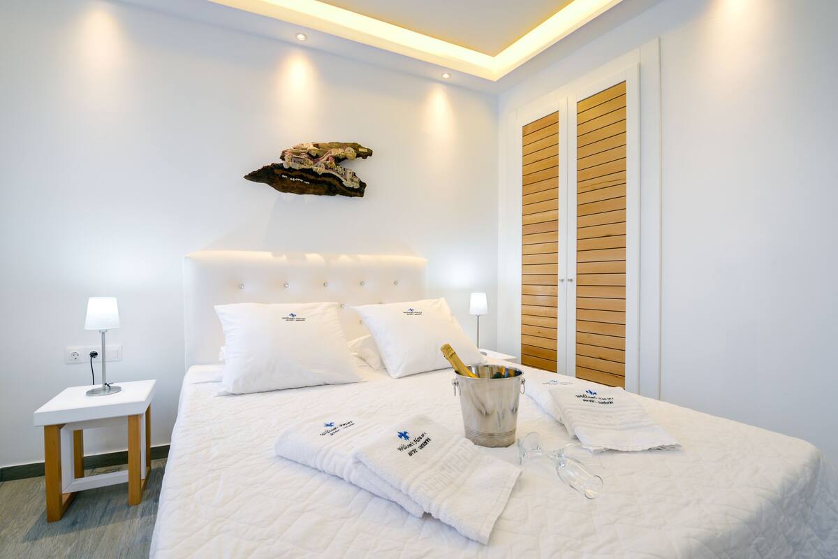 A hotel room with a neatly made bed, a small bedside table with a lamp, a bottle of champagne, and a bucket with glasses.