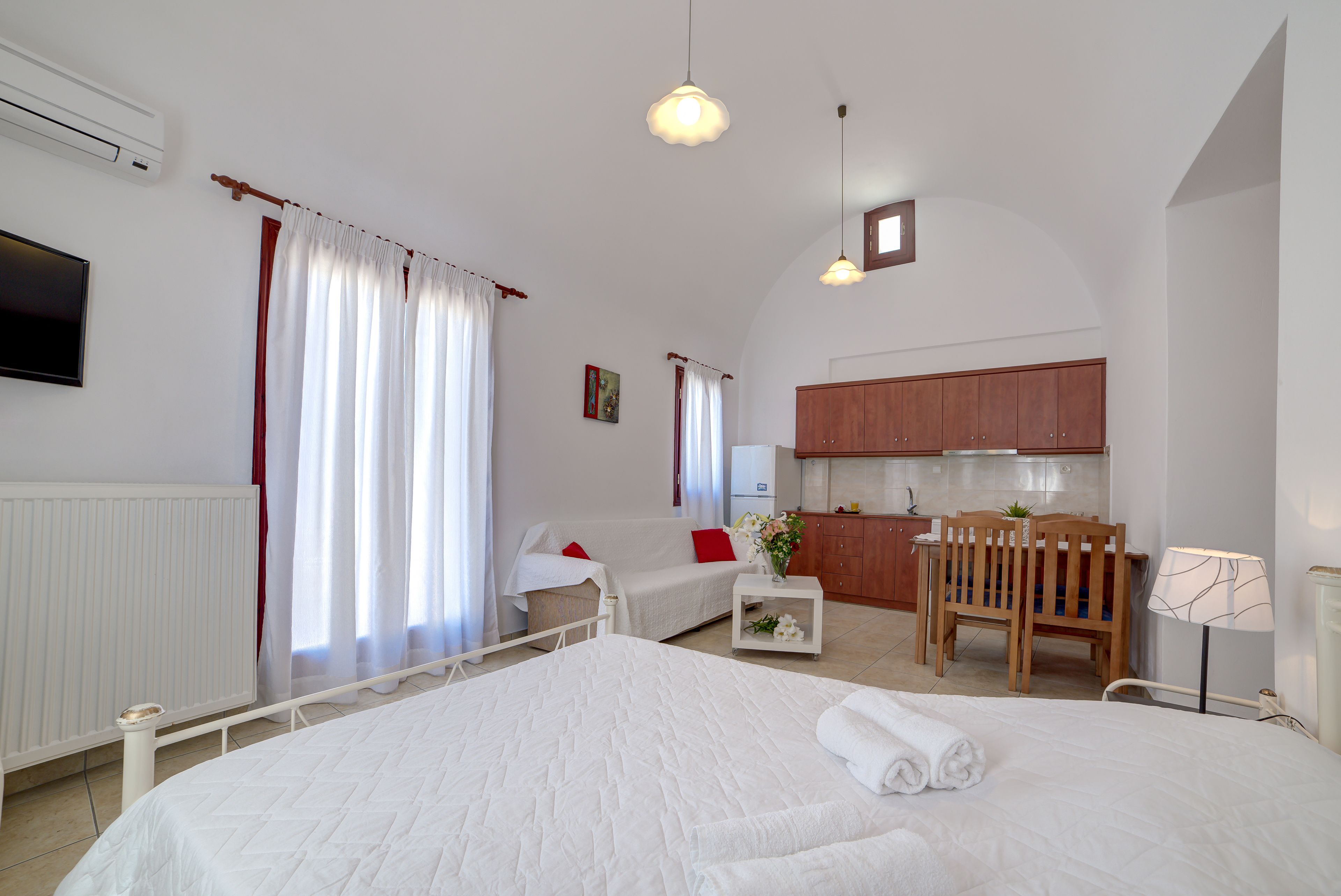 A bedroom with a large bed, a small desk, a chair, a sofa, and a kitchen area.