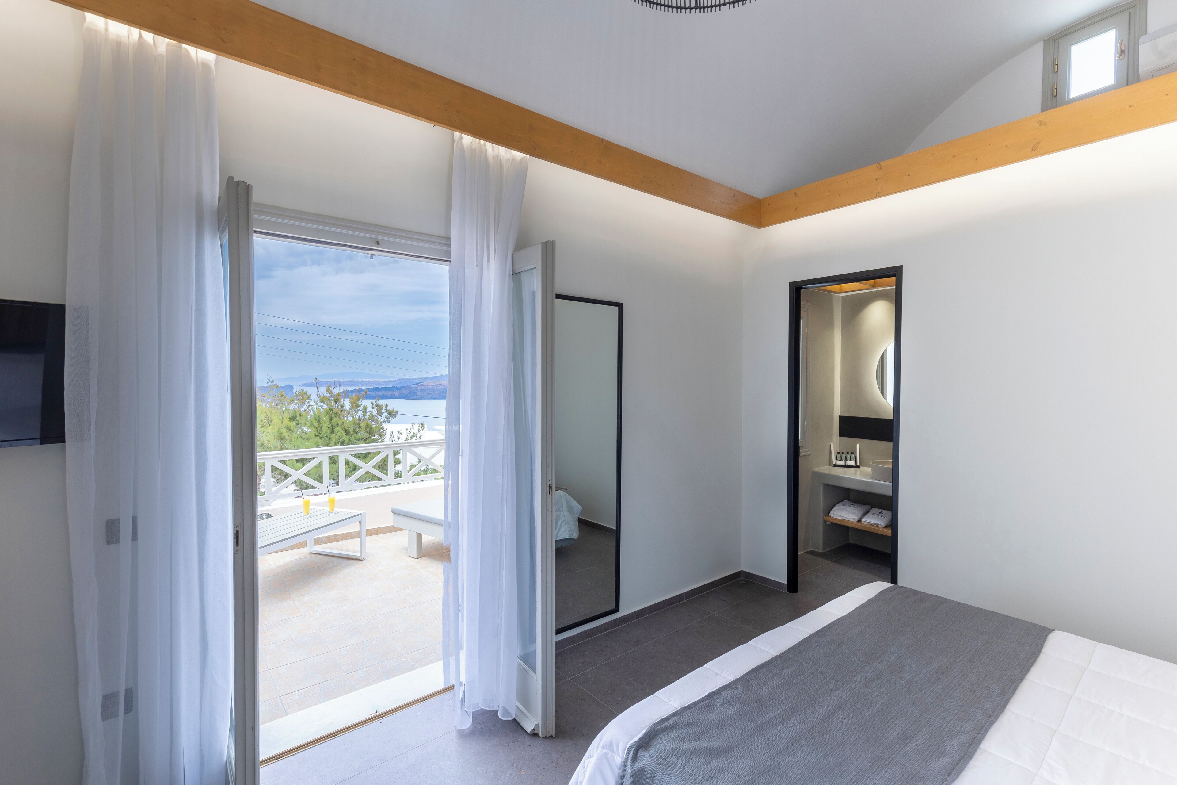 A bedroom with a bed, a large mirror, a wardrobe, and a sliding glass door leading to an outdoor area with a bench and a view of the sea.