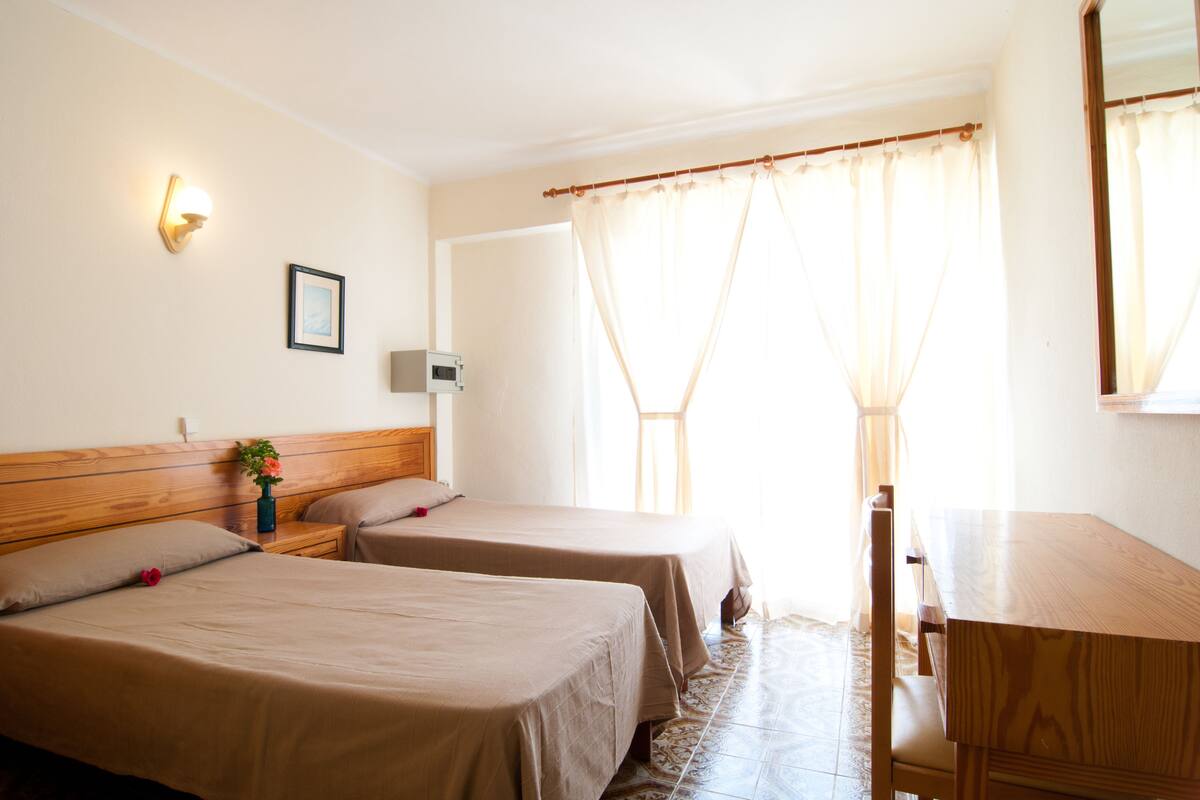 A hotel room with two single beds, a wooden desk, a chair, a nightstand with a lamp, and a framed picture on the wall.