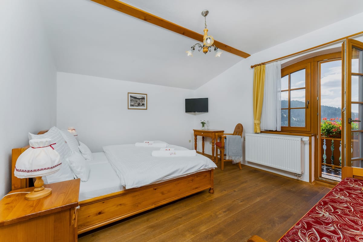A bedroom with a wooden bed, a bedside table, a small table with a chair, a TV mounted on the wall, and a window with curtains.