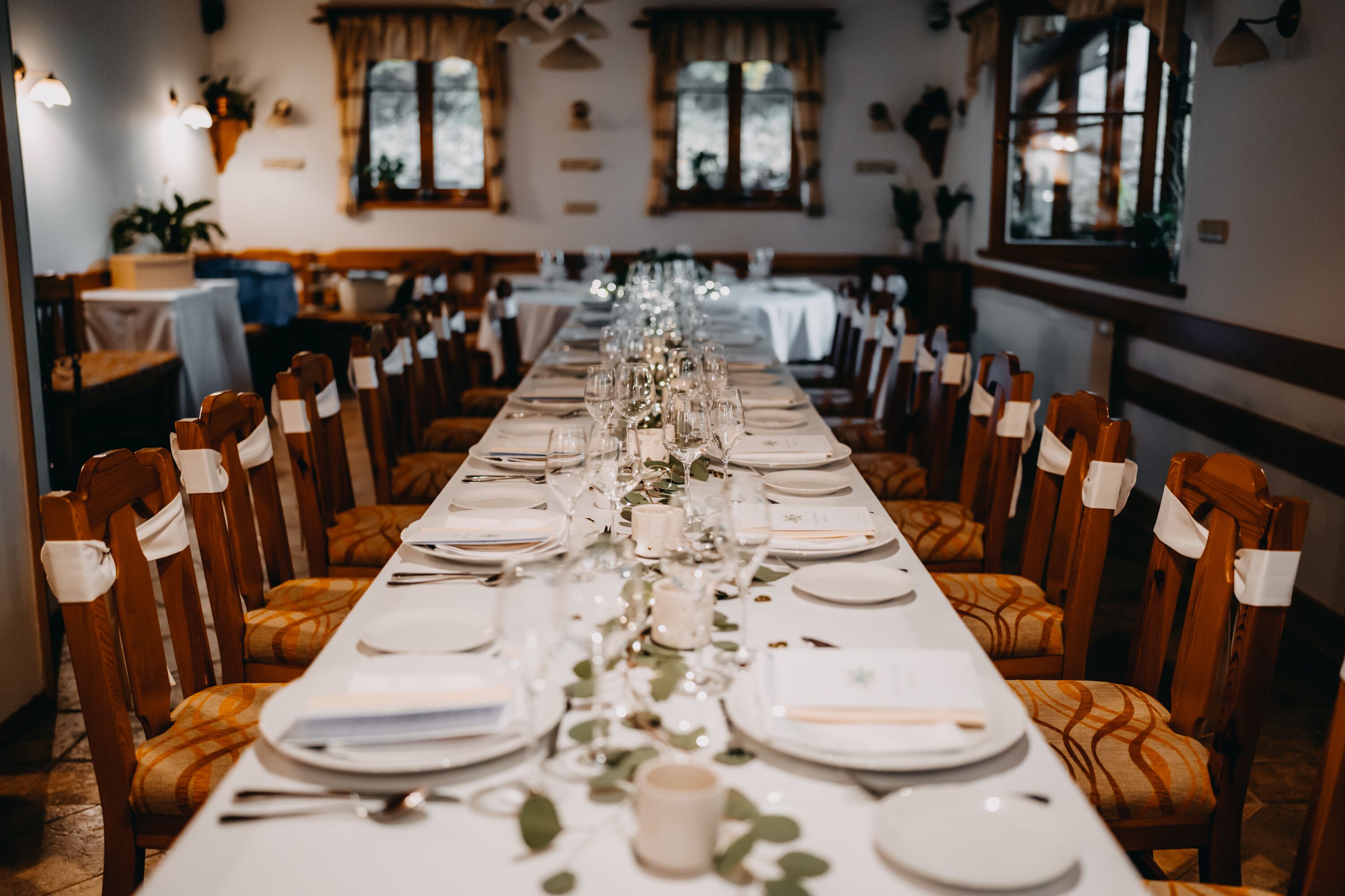 Una larga mesa de comedor dispuesta con sillas, platos y cubiertos, en una habitación con sillas de madera y ventanas.