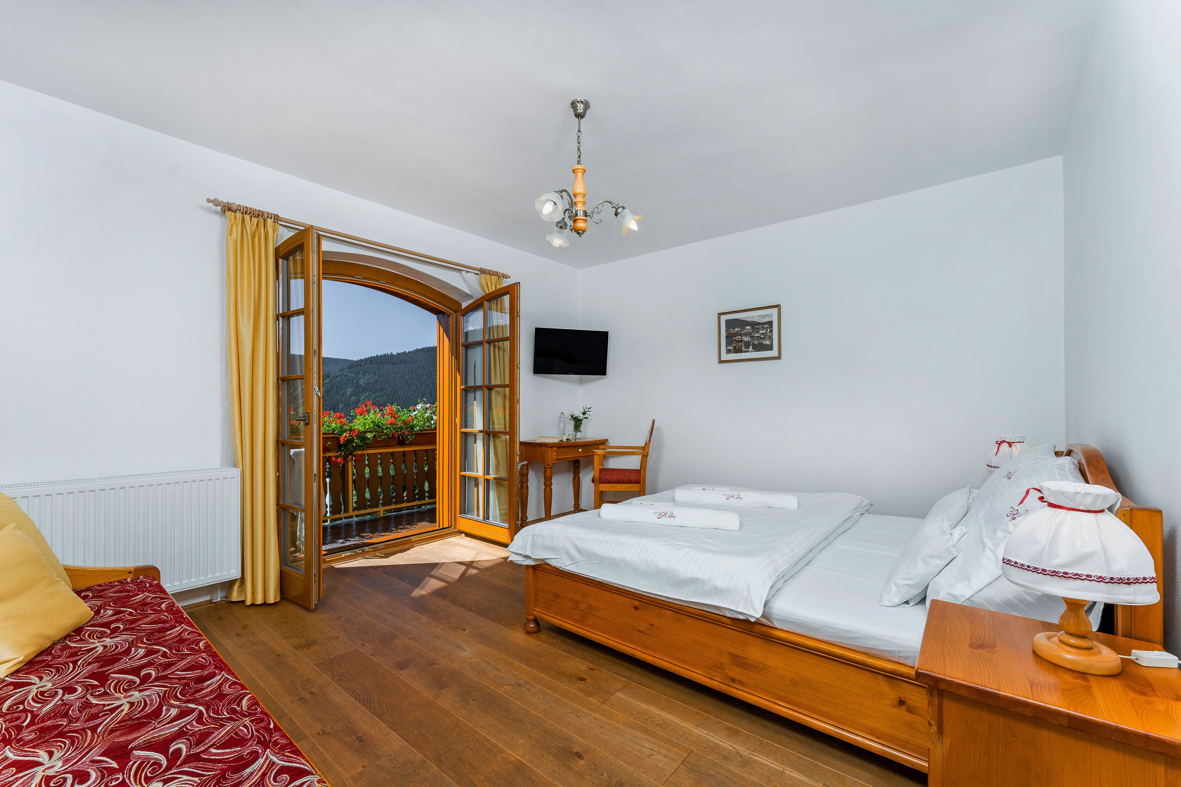 A bedroom with a wooden bed, a desk with a chair, a television on the wall, and a view through an open door.