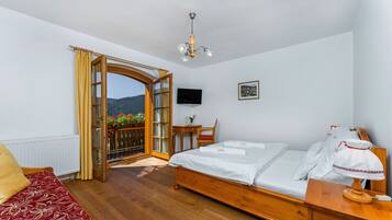 A bedroom with a wooden bed, a desk with a chair, a television on the wall, and a view through an open door.