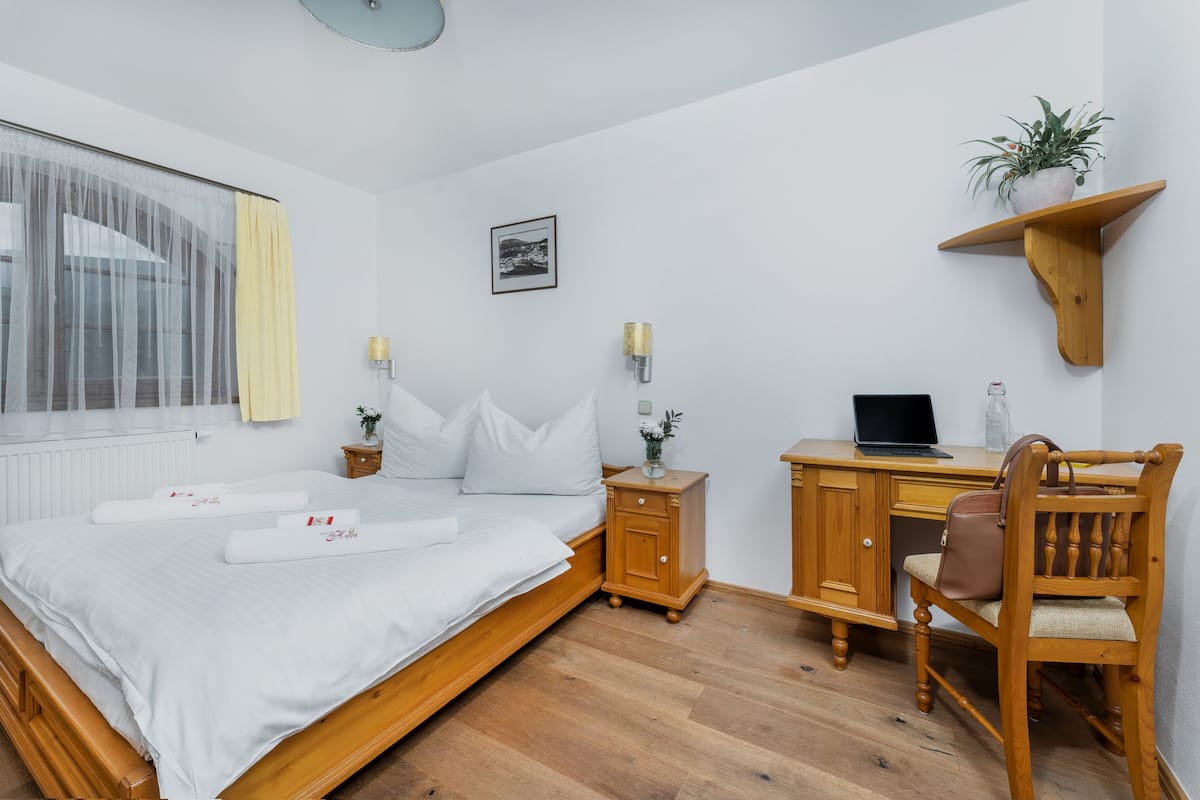 A bedroom with a bed, bedside tables, a desk, a chair, a shelf with a plant, and a picture on the wall.