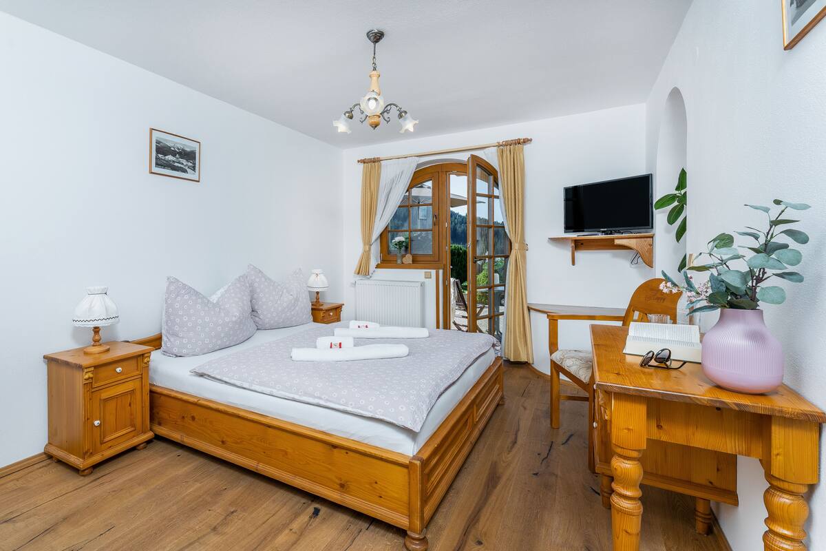 A bedroom with a wooden bed, a desk with a vase, a chair, a television, and a window with curtains.