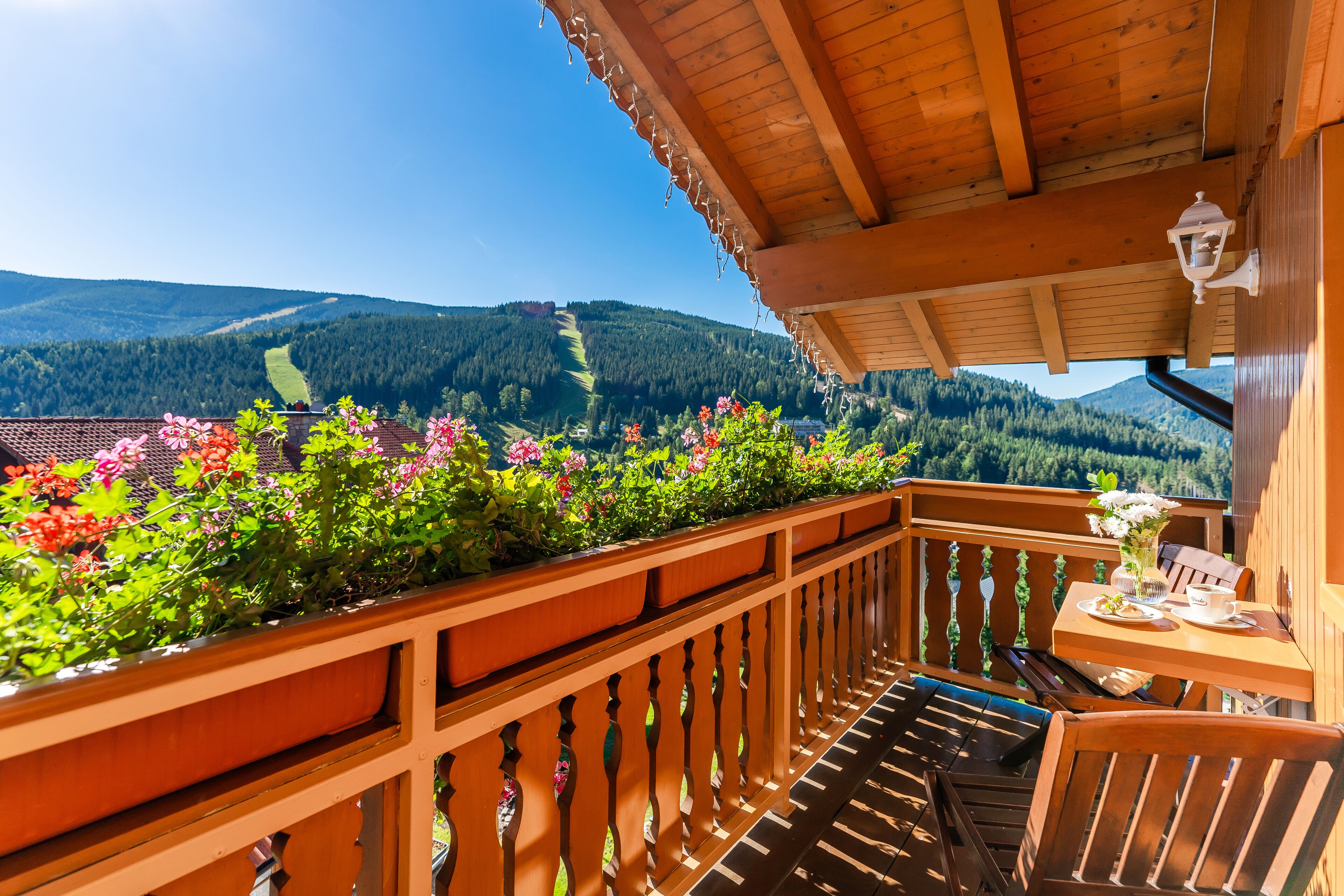 A wooden balcony with a view of mountains, a table with a chair, and a potted plant with flowers.