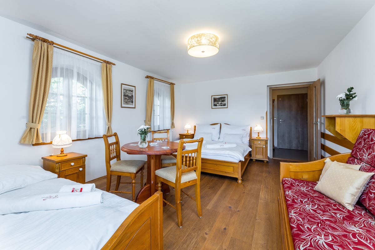 A hotel room with a wooden bed, a red sofa, a wooden dining table with chairs, a small round table with a lamp, and a door leading to a hallway.