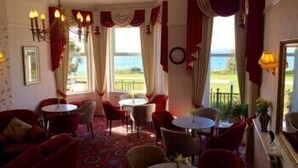 A cozy room with a patterned carpet, red upholstered chairs, and tables. There are large windows with curtains, a view of the outdoors, and a chandelier. - Hotel Riviera (Bournemouth)