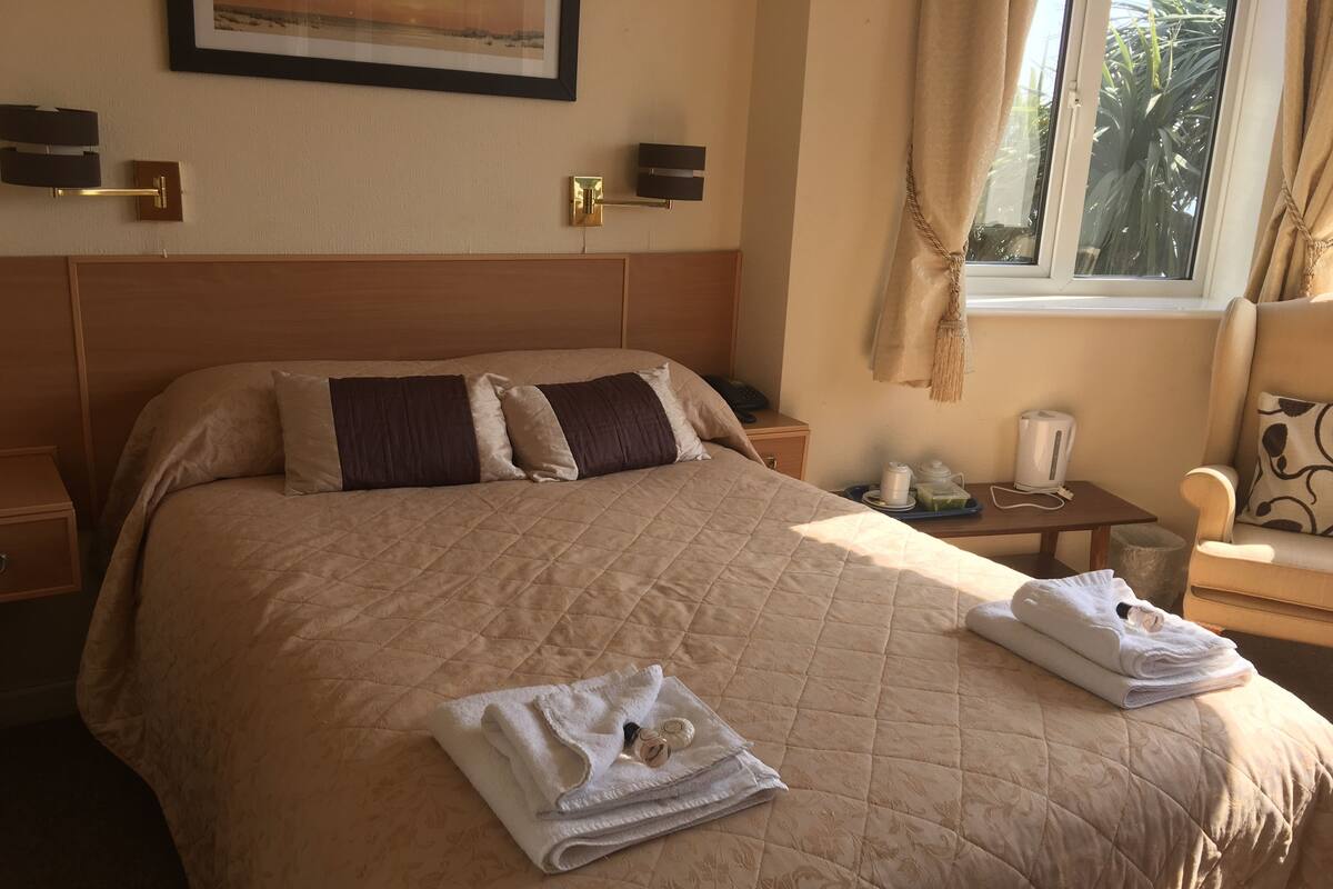 A hotel room with a large bed, a bedside table with a lamp, a small table with a kettle, and a window with curtains.