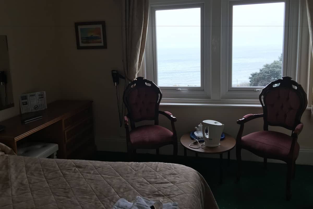 A hotel room with a bed, two chairs, a small table with a kettle, and a window with a view of the sea.