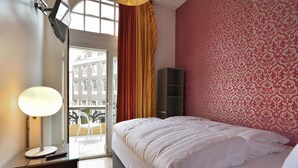 A hotel room with a bed, a desk with a lamp, a wardrobe, and a view of buildings through a window.