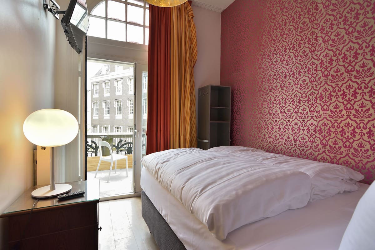 Habitación de hotel con cama, escritorio con lámpara, armario y vistas a edificios por la ventana.