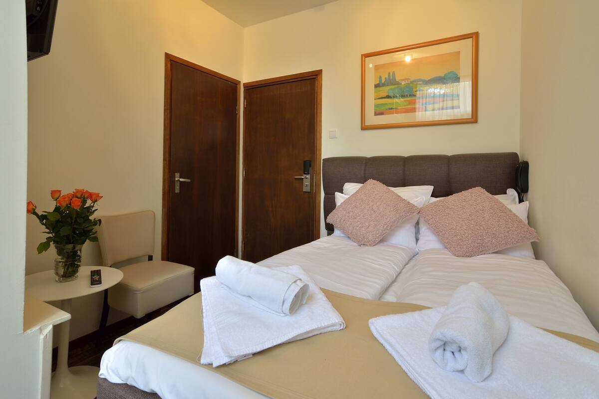 Habitación de hotel con una cama, dos puertas, una mesita con un florero y un cuadro en la pared.
