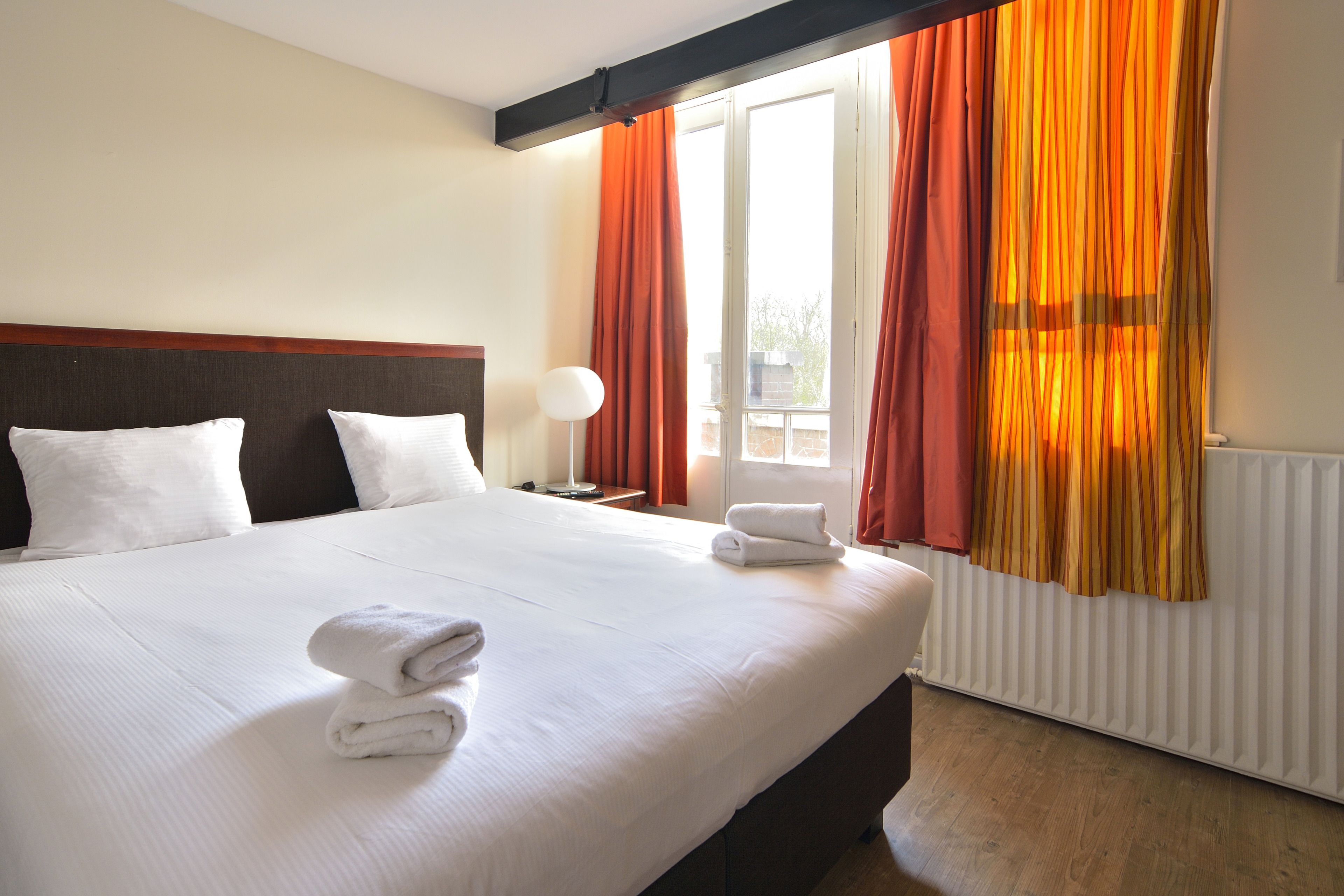 A hotel room with a large bed, white bedding, a nightstand with a lamp, and a window with orange curtains.
