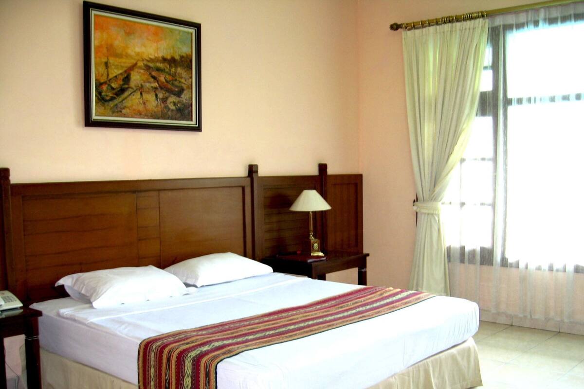 A hotel room with a wooden headboard, a bed with white linens, a bedside lamp, and a painting on the wall.