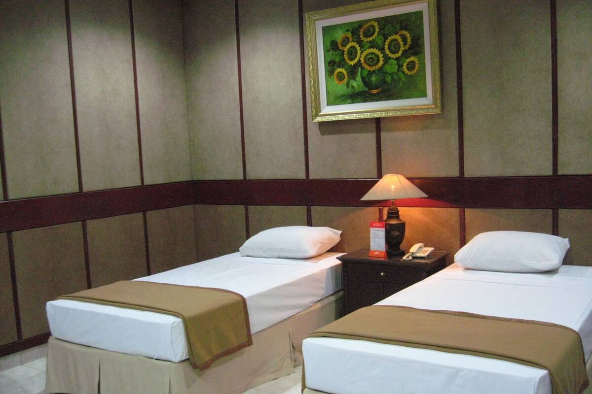 A hotel room with two single beds, a nightstand with a lamp, a framed painting on the wall, and a wooden headboard.