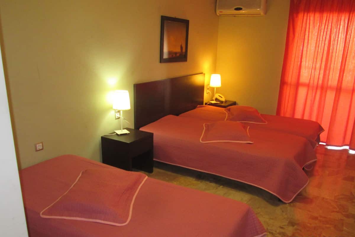 A hotel room with two beds, a nightstand with a lamp, a wall-mounted air conditioner, and a window with red curtains.
