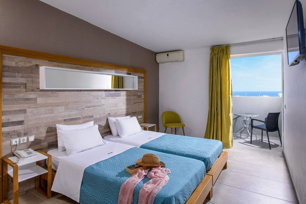 A hotel room with two beds, a wooden headboard, a small table with a phone, a chair, and a balcony with a table and chairs.