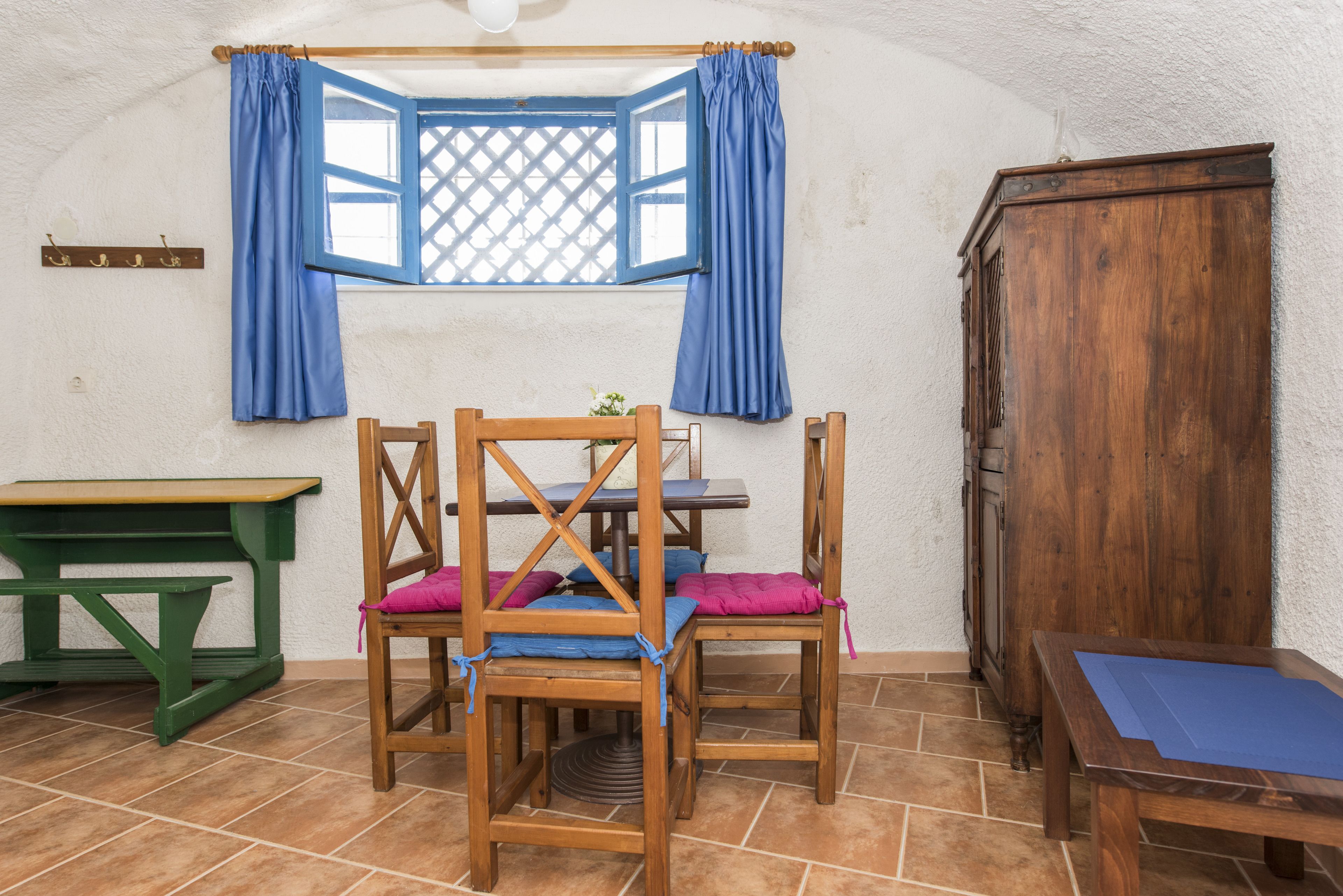 A room with a wooden table, two chairs with cushions, a wooden cabinet, and a window with blue curtains.