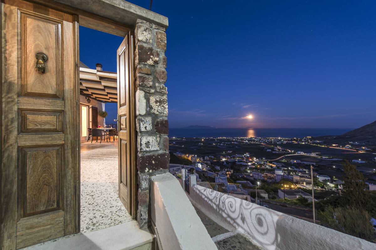 Una puerta de madera abierta que da a una habitación con pared de piedra, vista de un pueblo por la noche y una luna en el cielo.