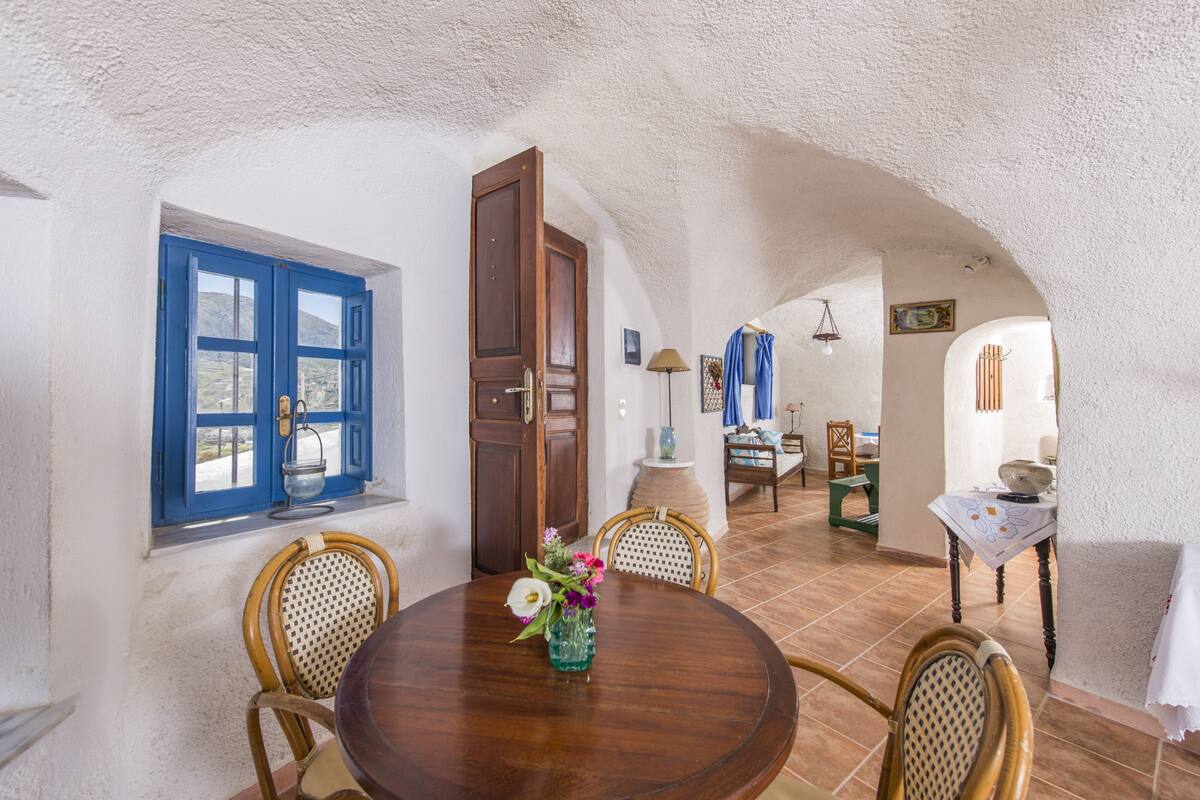 Un comedor con una mesa redonda de madera, sillas de mimbre y un jarrón de flores. Se aprecia una ventana con contraventanas azules y una puerta que da a otra habitación.